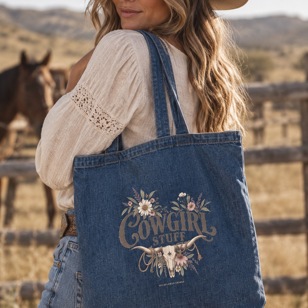 A woman outdoors wearing a denim tote bag over her shoulder. The tote bag has a floral and western-style graphic that reads 'Cowgirl Stuff' with a longhorn illustration.