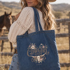 A woman outdoors wearing a denim tote bag over her shoulder. The tote bag has a floral and western-style graphic that reads 'Cowgirl Stuff' with a longhorn illustration.