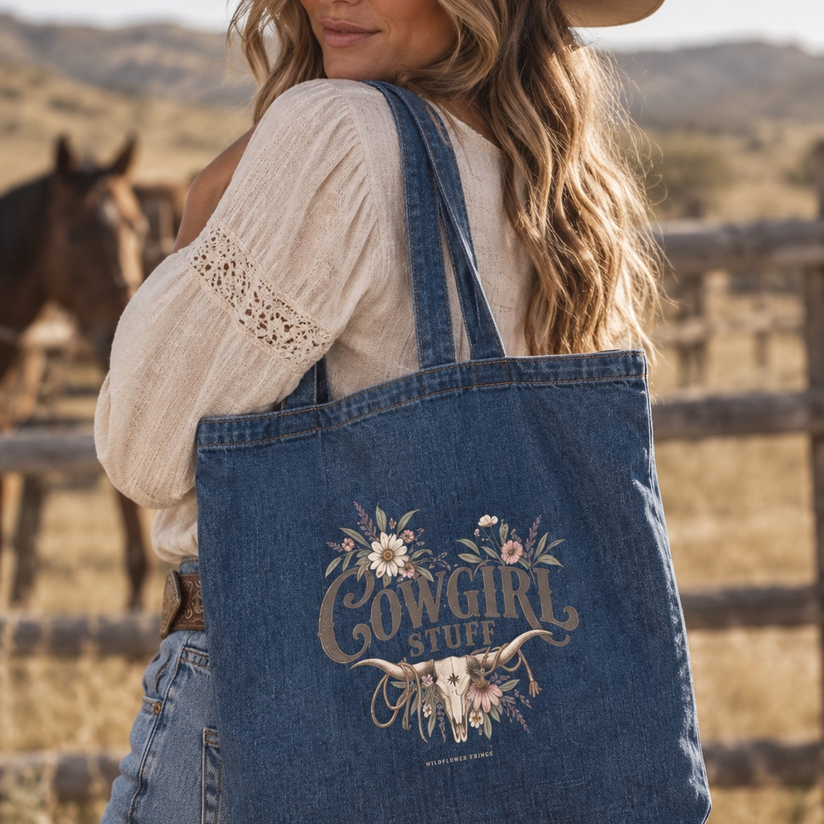 A woman outdoors wearing a denim tote bag over her shoulder. The tote bag has a floral and western-style graphic that reads 'Cowgirl Stuff' with a longhorn illustration.