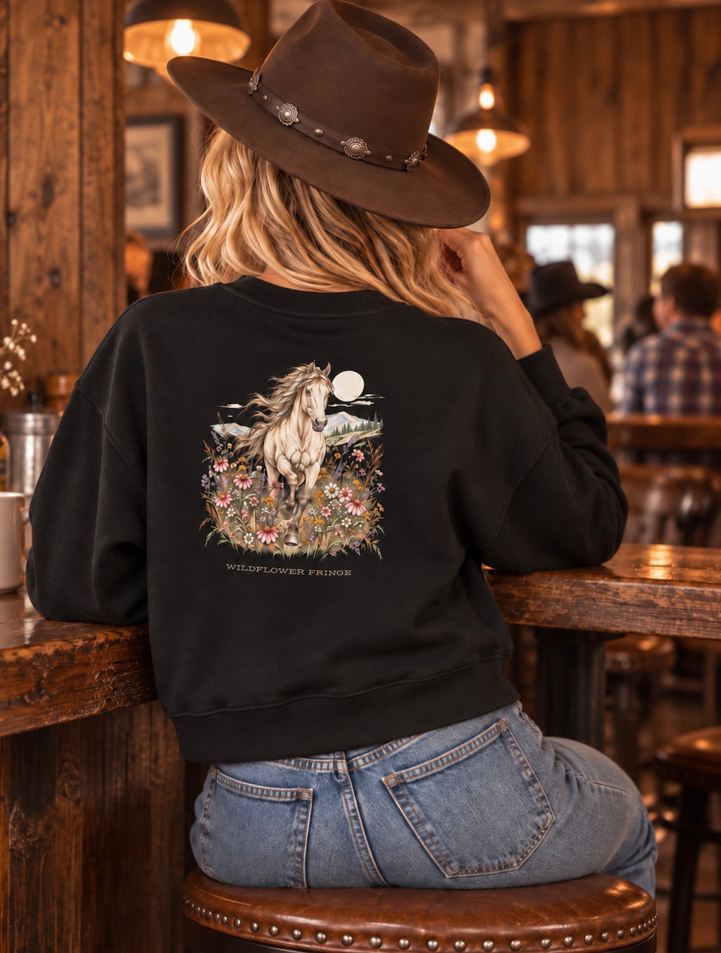 A woman wearing a black crewneck sweatshirt with a detailed western-style horse graphic on the back, sitting on a wooden stool inside a rustic bar or restaurant.