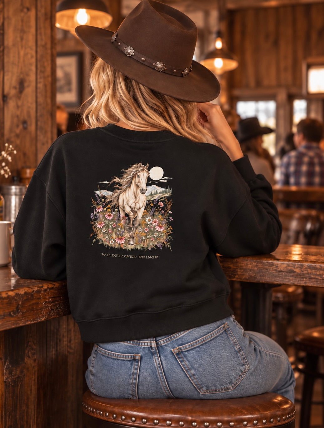 A woman wearing a black crewneck sweatshirt with a detailed western-style horse graphic on the back, sitting on a wooden stool inside a rustic bar or restaurant.