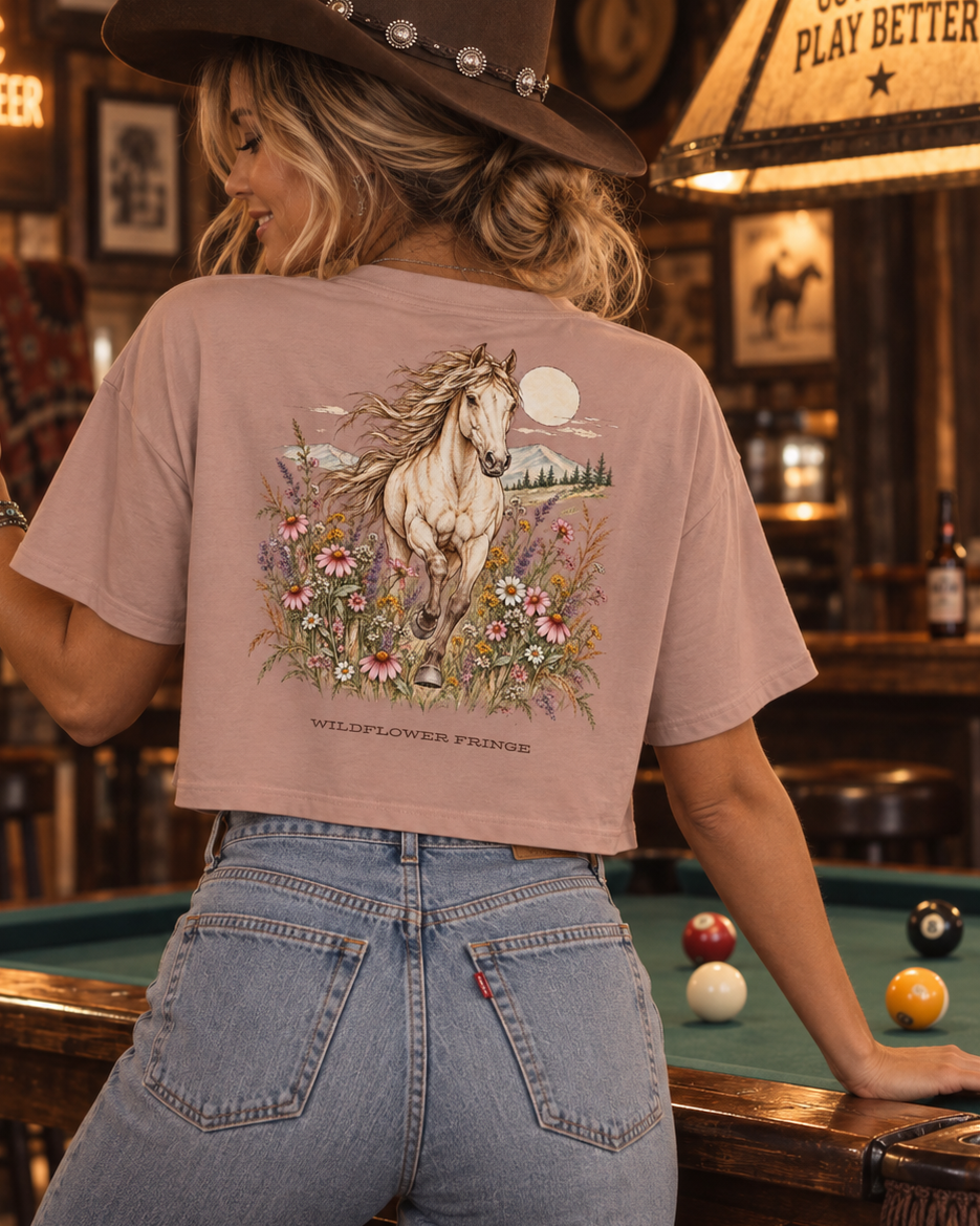 A woman in a cowboy hat and jeans stands in a rustic bar, seen from behind, wearing a dusty rose graphic t-shirt with a detailed western horse print on the back.