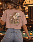 A woman in a cowboy hat and jeans stands in a rustic bar, seen from behind, wearing a dusty rose graphic t-shirt with a detailed western horse print on the back.