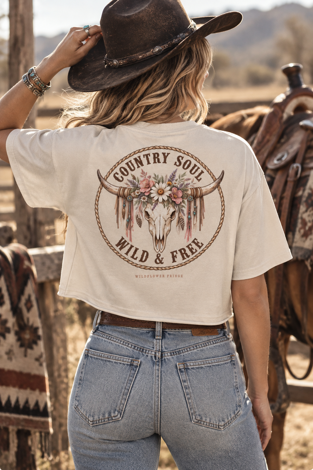 A woman from behind wearing a cream-colored cropped t-shirt with a large western-themed graphic on the back, paired with light-wash jeans and a dark brown cowboy hat, set in a rustic outdoor ranch environment with a horse in the background.