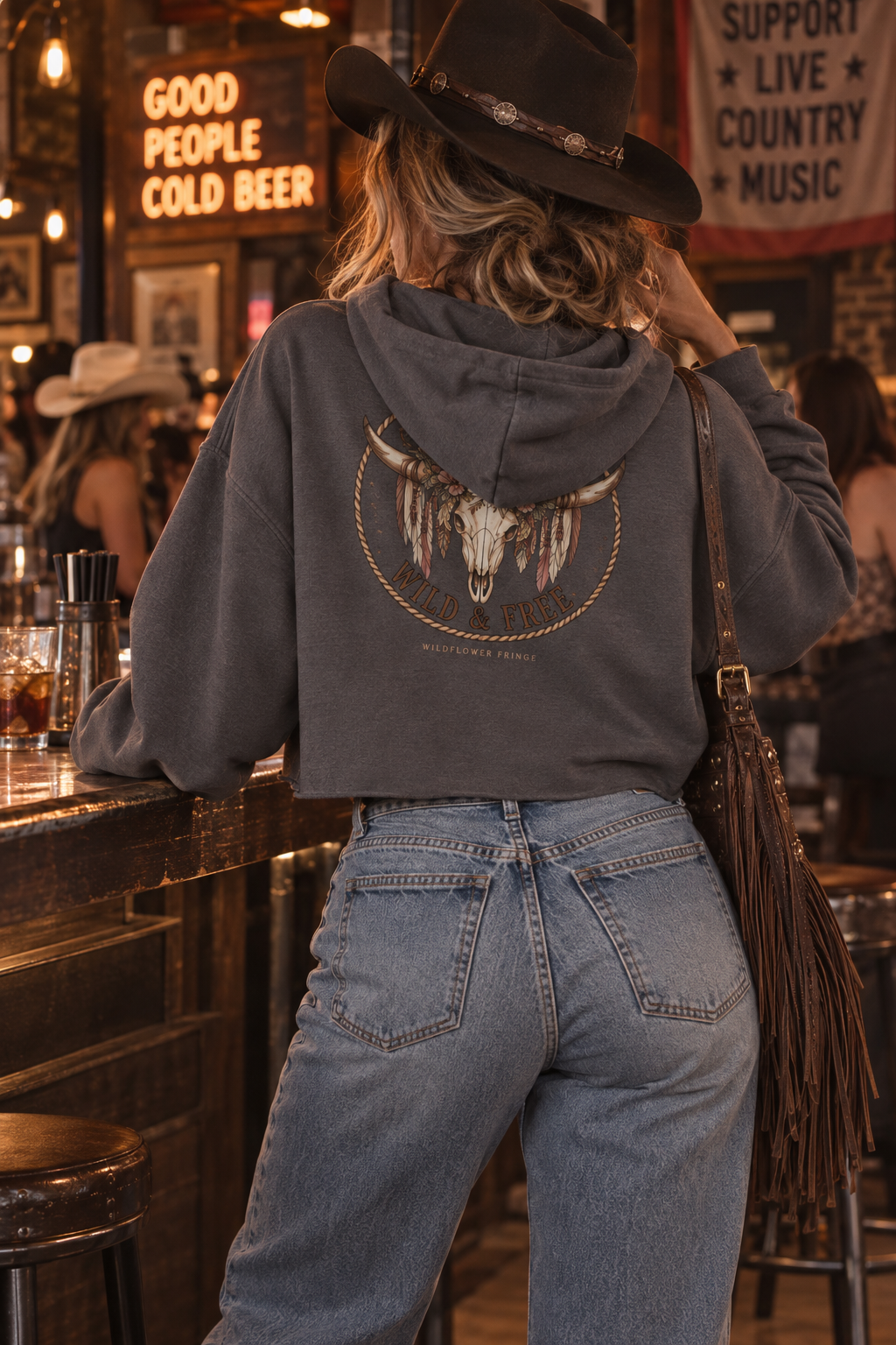 A woman wearing a dark gray hooded sweatshirt with a large Native American skull graphic on the back, paired with light blue jeans and a black cowboy hat, standing at a bar in a rustic, warmly lit environment.
