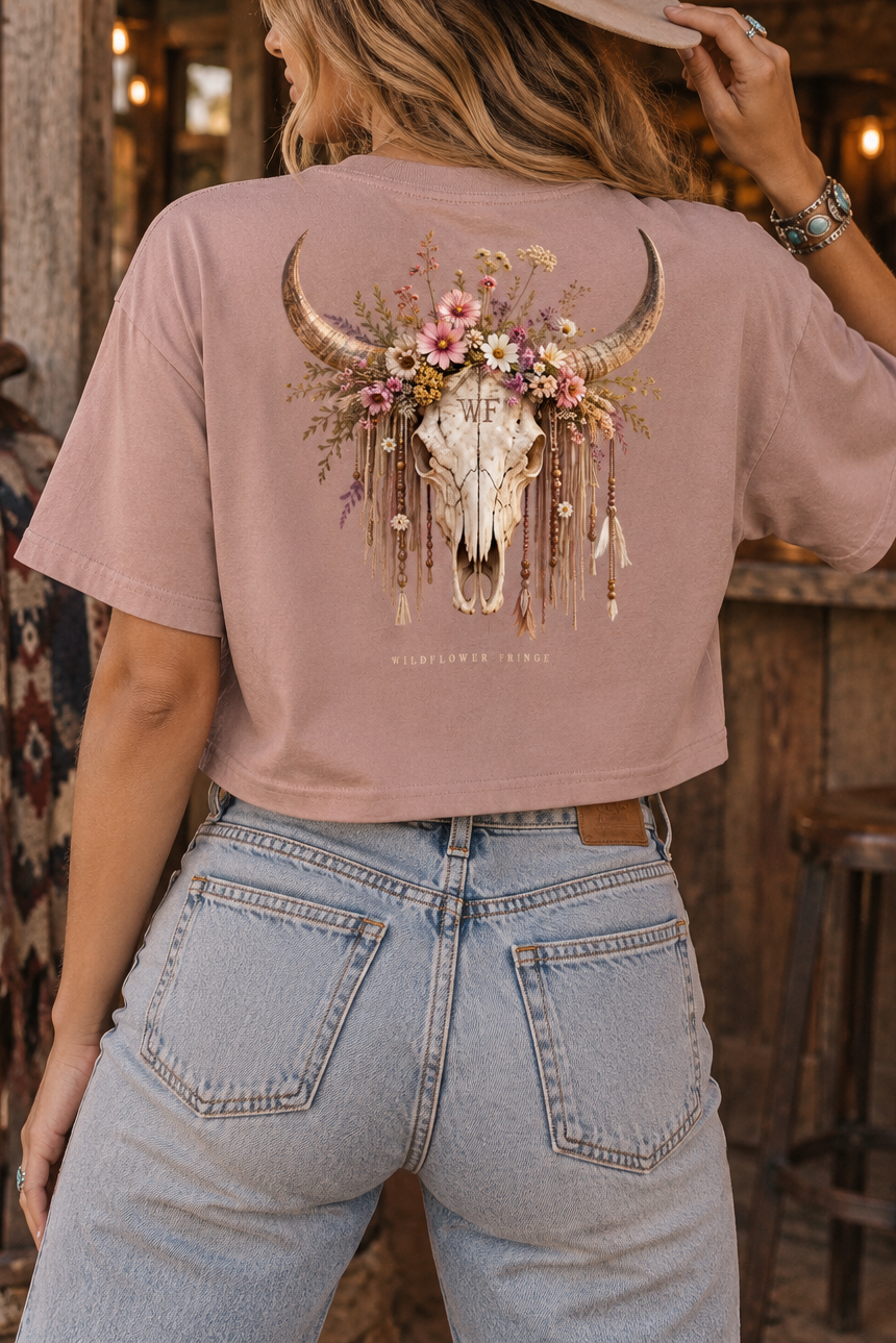 A woman wearing a dusty rose cropped t-shirt with a large bohemian-style bull skull and floral graphic on the back, paired with light-wash jeans and a tan cowboy hat, standing in a rustic western-themed bar.