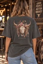 A woman viewed from behind wearing a dark charcoal gray t-shirt with a large graphic of a decorated skull and floral horns, paired with light blue jeans in a rustic bar setting.