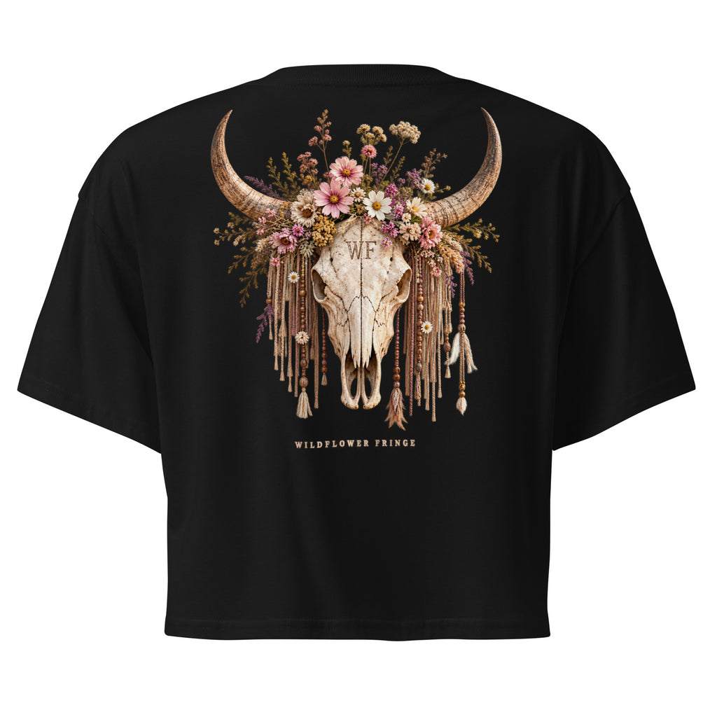 Black short-sleeve crop top t-shirt showing the back graphic, which features a decorated animal skull with curved horns, a floral crown, and dangling tassels. The text 'WILDFLOWER PRINCE' is printed below the graphic.