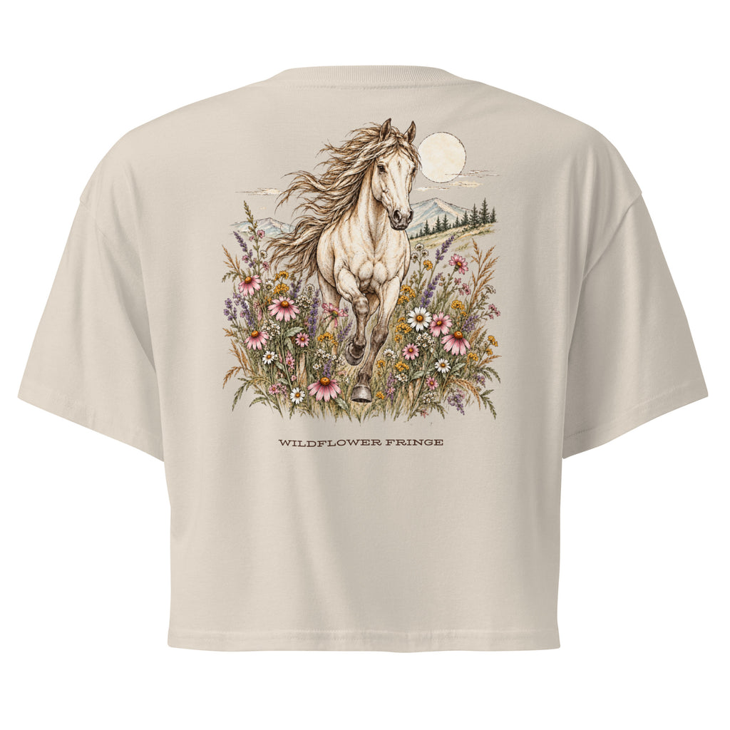 A beige short-sleeve crop top shown from the back, featuring a large graphic print of a white horse running through colorful wildflowers under a full moon, with the text 'WILDFLOWER FRINGE' printed below.