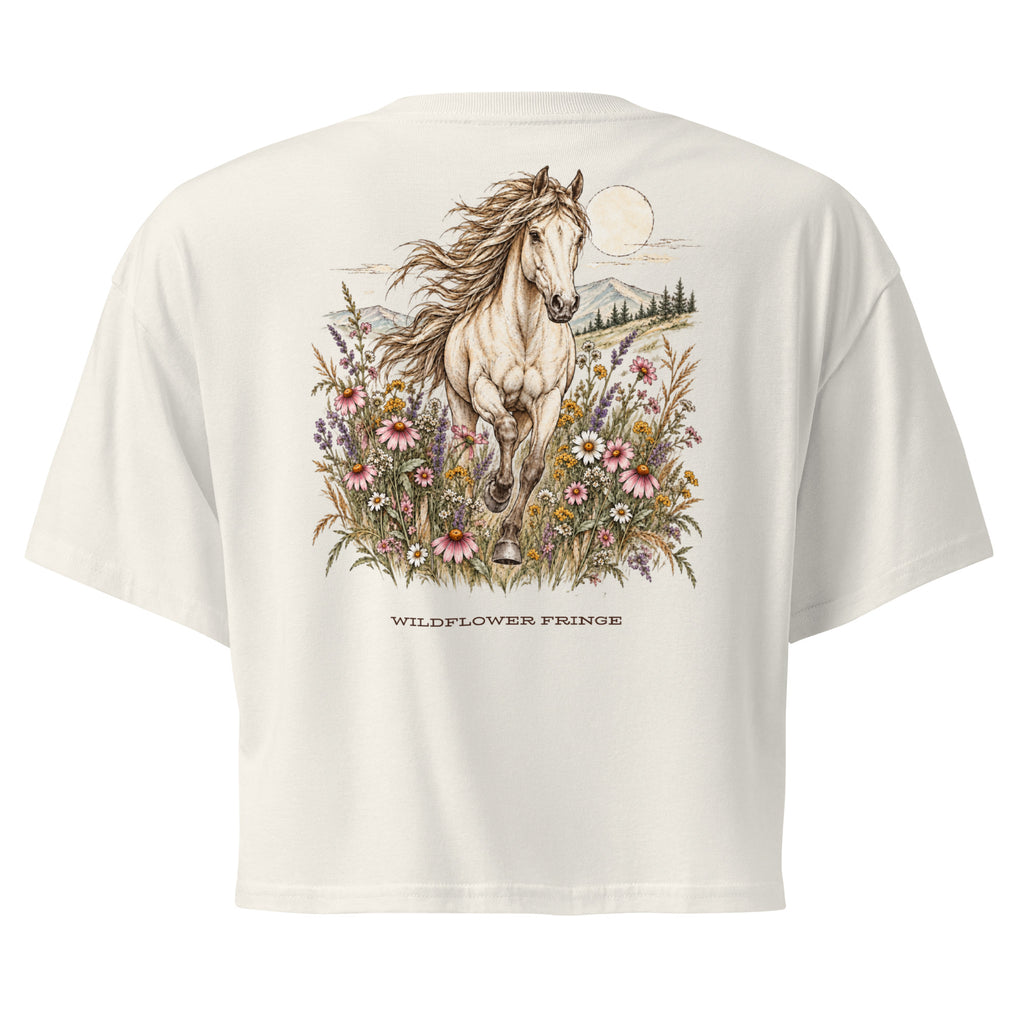 An off-white short-sleeve crop top shown from the back, featuring a large graphic of a white horse running through colorful wildflowers under a full moon, with the text 'WILDFLOWER FRINGE' printed below.