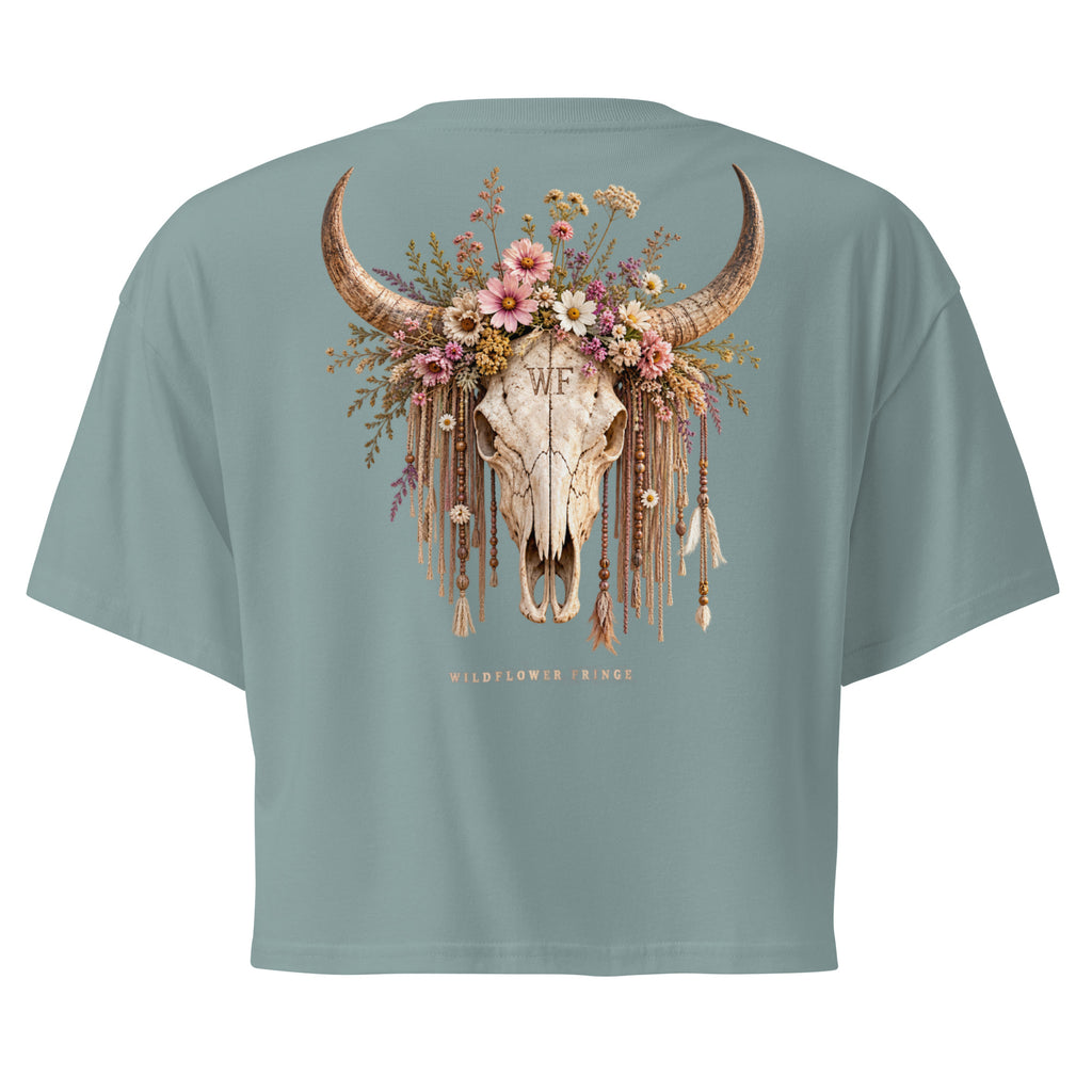 A sage green short-sleeve crop top shown from the back, featuring a large graphic of a decorated skull with long horns, a floral crown, and dangling tassels. The text 'WF' is printed on the skull, and 'WILDFLOWER TRIBE' is printed below.
